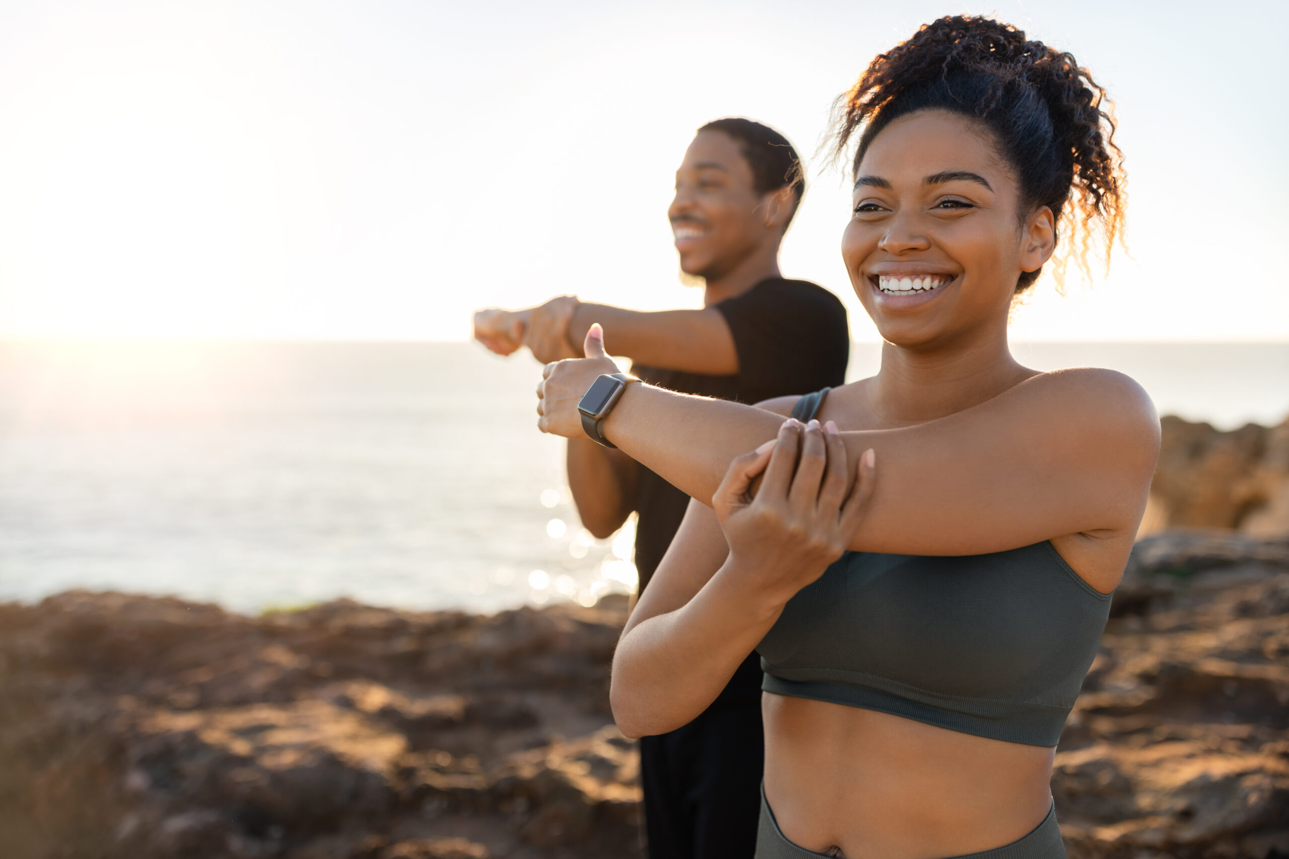 Smiling millennial african american lady and male in sportswear doing arm stretching, enjoy morning workout on ocean beach at sunrise. Body care and health care together, weight loss outdoor Elysian Medspa & Rejuvenation Center in Indianapolis